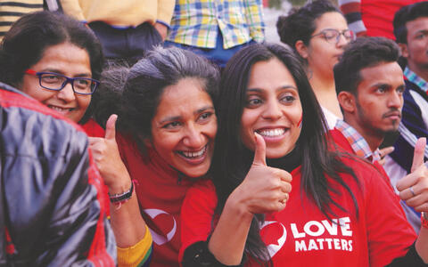 Drei Frauen mit roten Shirts mit der Aufschrift "Love Matters" stehen in einer Menschengruppe und halten die Daumen nach oben
