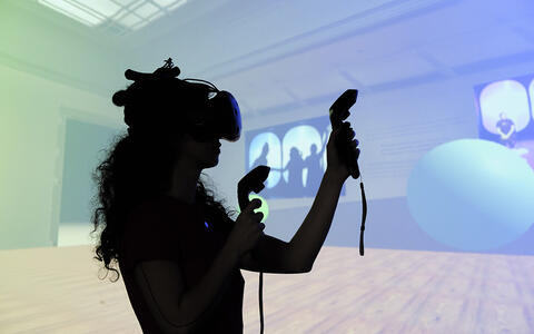 Studentin steht mit einer VR-Brille und Controllern vor einer farbigen Wand