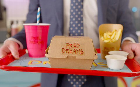 Das Bild zeigt einen Mann in einem blauen Anzug, der ein Tablett mit einer braunen Papierschachtel mit der Aufschrift „Fried Dreams“ und einem roten Tablett hält. Das Tablett enthält auch einen rosa Becher mit einem gestreiften Strohhalm, einen kleinen weißen Becher und einen gelben Becher mit Pommes frites. Der Hintergrund ist unscharf mit Küchen- und Essensgegenständen.