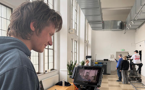 Eine Gruppe Studierender filmt im Hintergrund eine Szene. Im Vordergrund schaut ein Student auf einen Bildschirm mit dem Kamerabild