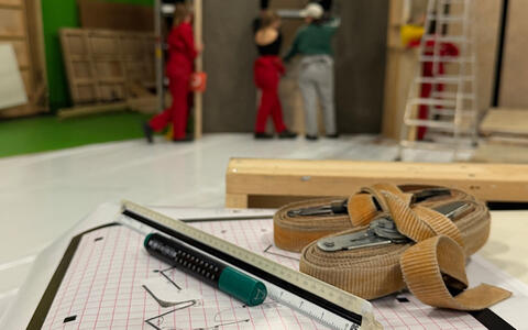 Ein Stift, ein Lineal und Spanngurte liegen auf einem Bauplan. Im Hintergrund sind ifs-Studierende beim Aufbau von Wänden