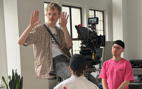 Ein Student sitzt mit nach oben gerichteten Händen auf einem Dolly, hinter ihm die Kamera, die auf einen weiteren Studenten gerichtet ist. Vor ihm steht eine weitere Studentin