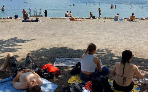 Fotografie eines sonnigen Strandes mit einer bunten Menschenmenge, die das Wasser genießt. Im Vordergrund sitzen zwei Frauen auf Handtüchern, während ein Mann ohne Hemd in der Nähe liegt. Berge und ein teilweise bewölkter Himmel bilden den Hintergrund.