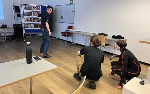 Auf diesem Foto steht ein junger Mann in schwarzem Hemd und Jeans in einem minimalistischen Klassenzimmer mit Holzboden, weißen Wänden und einfachen Möbeln. Zwei Frauen in schwarzer und roter Sportkleidung hocken vor ihm. Eine Wasserflasche und ein Laptop stehen auf einem Tisch.