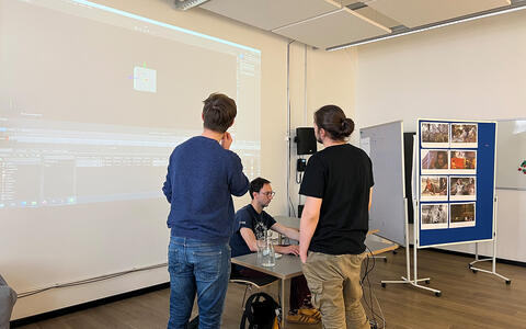 Foto von drei jungen Männern in einem modernen, hellen Konferenzraum. Zwei Männer in Freizeitkleidung stehen an einem Projektor, der einen Bildschirm mit Text und Bildern zeigt. Ein dritter Mann sitzt an einem Tisch, trägt ein schwarzes T-Shirt und eine Brille und arbeitet an einem Laptop. Der Raum ist mit weißen Wänden, Holzfußboden und einer blauen Pinnwand mit Fotos ausgestattet.
