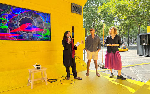 Die MA Digital Narratives Studierenden, Amna Tariq, Haydar Tastan, and Moheman Iqbal präsentieren ihr Project. Sie stehen auf einem gelben Boden vor einer gelben Wand in der ein Fernseher eingelassen ist. Auf dem Fernseher sieht man bunte Formen und auf dem Boden ist ein kleiner Hocker auf dem eine VR-Brille liegt.