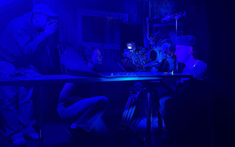 Dunkle Umgebung mit leicht blauem Licht. Eine junger Mann sitzt an einem Tisch, auf ihn ist eine Filmkamera gerichtet. Daahinter hockt eine Frau auf dem Boden. Zwei Sudierende filmen ihn mit anderen Kameras