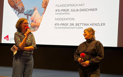 Zwei Frauen mit Mikrofon in der Hand stehen in einem Kinosaal vor einer Leinwand.
