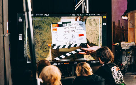 Eine Filmklappe auf einem großen Bildschirm. Davor sitzt eine Gruppe Studierender