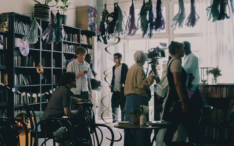 Eine Filmcrew am für eine Party dekorierten Set in einem Raum mit kleinen Tischen und Stühlen und Bücherregalen an der Wand.