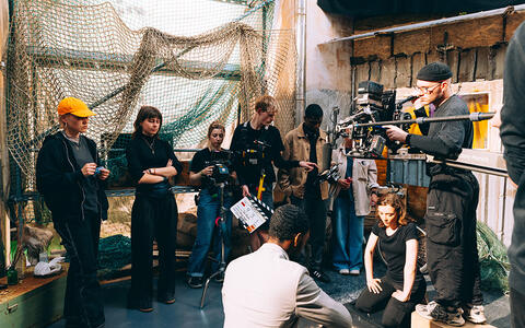 Eine Frau und ein Mann sitzen am Boden. Ein Kameramann filmt die Szene von oben. Eine Filmcrew steht um sie herum