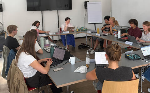Eine Gruppe sitzt an Tischen in einem Seminarraum, einige haben einen Laptop vor sich stehen