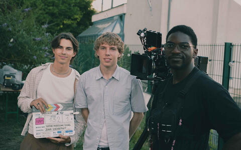Drei junge Männer. Einer hält eine Filmklappe in der Hand, zwischen den anderen beiden steht eine Filmkamera.