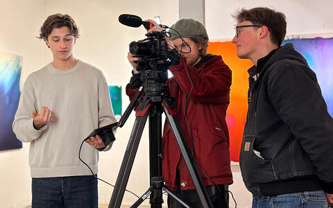 Ein Filmteam steht in einer Galerie hinter einer Kamera und beobachtet das Kamerabild