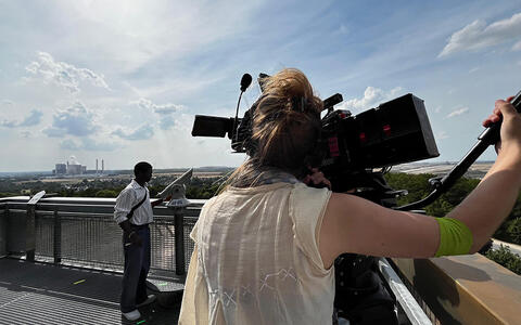 Eine junge Frau filmt einen jungen Mann mit einer Filmkamera auf einem Aussichtsturm. Im Hintergrund sieht man Häuser und Industrieschornsteine.