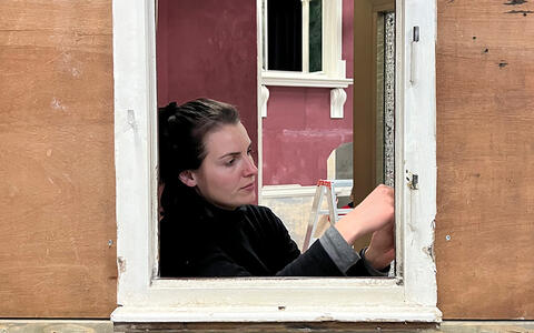 Eine Studentin ist in einem Fesnterahmen zu sehen und arbeitet an diesem. Im Hintergrund ist ein weiteres Fenster zu sehen untem dem eine Leiter steht.