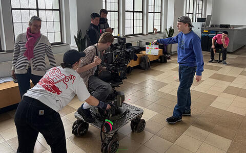 Zwei Studierenden areiten mit dem Dolly und der Kamera, die auf einen Studenten gerichtet ist. Ein weiterer Student steht dazwsichen und hält eine Filmklappe in die Kamera