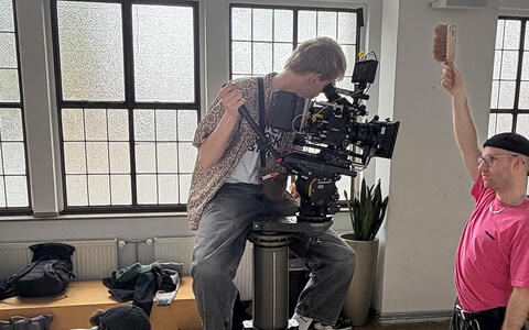 Ein Student sitzt auf dem Dolly und schaut durch die Kamera. Ein anderer Student steht vor der Kamera und hält einen Handfeger hoch