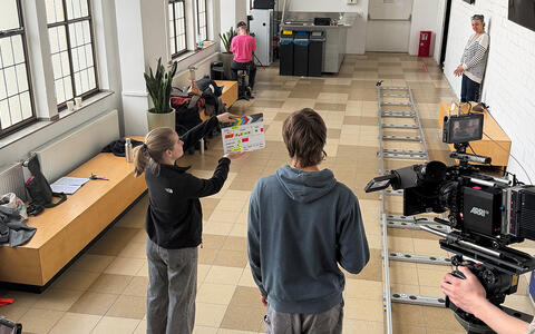 Zwei Studierende im Vordergrund, die Stundentin hält eine Filmklappe auf die eine Kamera gerichtet ist. Rechts liegen Schienen auf dem Boden die weiter in den Raum führen. Weiter hinten steht die Dozentin und schaut zu