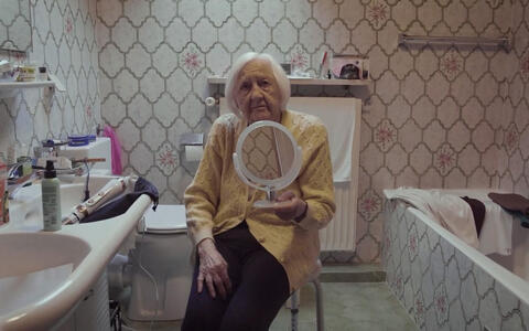 Eine alte Frau sitzt auf einem Hocker in einem Badezimmer mit einem Handspiegel in der Hand