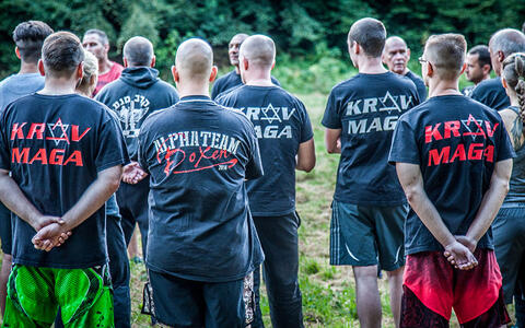 Foto einer Gruppe überwiegend weißer Männer, die schwarze T-Shirts mit der Aufschrift »KRAV MAGA« tragen und im Freien auf einem grasbewachsenen Gelände mit einem dichten Wald im Hintergrund stehen.