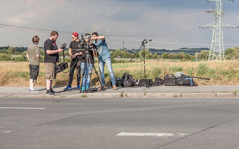 Foto von fünft Männern, die eine Szene an einer Landstraße mit Stromleitungen und einem hohen Strommast im Hintergrund filmen. Sie verwenden eine professionelle Videoausrüstung mit Kameras, Licht und Rucksäcken, die auf dem Grünstreifen verstreut sind.