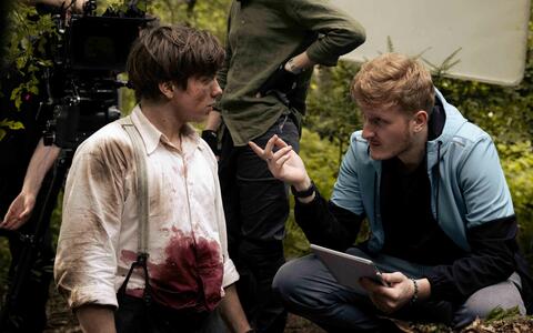 Still »Die letzte Mine« ifs filmschule köln Ein Crewmitglied ist in der Hocke, hält ein tablet in der Hand und spricht mit einem Schauspieler. Er hat ein weißes Hemd an, auf dem unechte Blutflecken zu sehen sind, wie von einer Schusswunde.