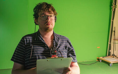 Am Set von »Dünn ist die Haut« im ifs-Studio: Ein Crewmitglied steht mit einem Klemmbrett und Headset vor einer Greenscreenwand.