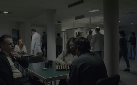 Ein geschäftiger, düster beleuchteter Empfangsbereich in einer Einrichtung. Drei Männer sitzen im Vordergrund an einem Tisch, von denen zwei Schach spielen.