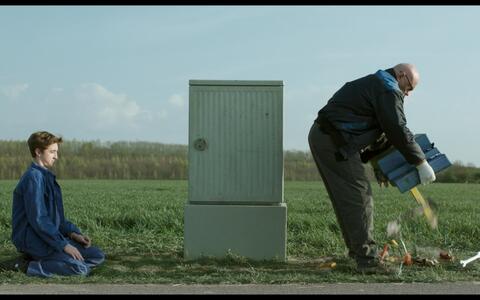 Auf einem offenen Feld am Tag reißt ein Mann Geräte aus dem Boden, während ein junger Mann auf dem Gras kniet und ihn beobachtet.