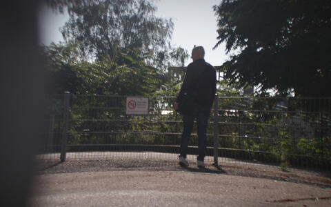 Ein Mann steht vor einem kleinen Gitterzaun und wird heimlich von hinten gefilmt.