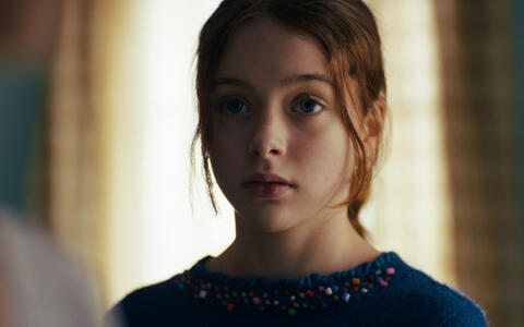 Ein Mädchen mit rötlichen Haaren und einem blauen Pullover steht vor einem Fenster