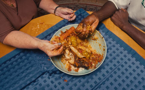 Ein Teller mit Essen von oben. Eine helhäutige Person und eine dunkelhäutige Person essen mit den Händen