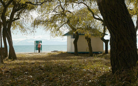Das Still zeigt Margarita Jumascheva, die an einer Küste steht. Im Hintergrund sind ruhige blaue Gewässer und ferne Berge unter einem teilweise bewölkten Himmel zu sehen.