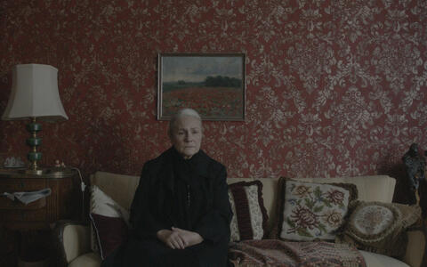 Eine alte Frau sitzt mit traurigem Blick ganz in schwarz gekleidet auf einem alten Sofa