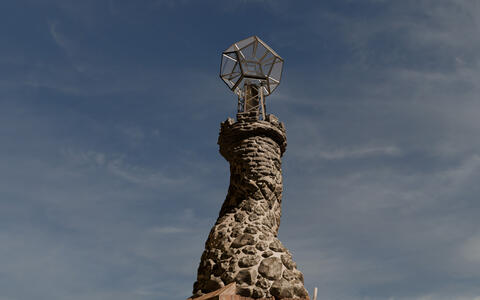 Das Foto zeigt einen Turm mit einer großen, komplizierten, metallischen, geometrischen Struktur auf der Spitze. Der Turm selbst ist aus rauen, grauen Steinen gebaut. Der Himmel darüber ist blau mit vereinzelten Wolken.