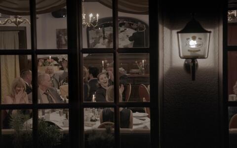 Die Filmszene wurde von außen durch ein Fenster in einen großen Festsaal in der Nacht aufgenommen. Drinnen sind viele gut gekleidete Leute. Die Stimmung ist fröhlich und festlich.