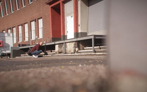 Ein Mann hockt vor einer Fabrik vor einer Laderampe und wird heimlich gefilmt.