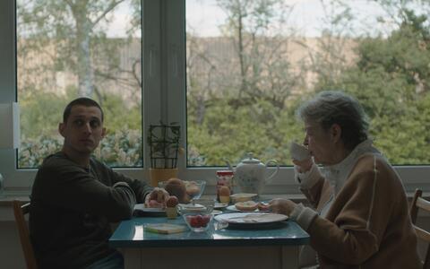 Ein junger Mann und eine ältere Frau sitzen sich an einem kleinen Tisch gegenüber und frühstücken. Die Frau nippt an ihrem Tee, während der Mann nachdenklich zur Seite schaut.