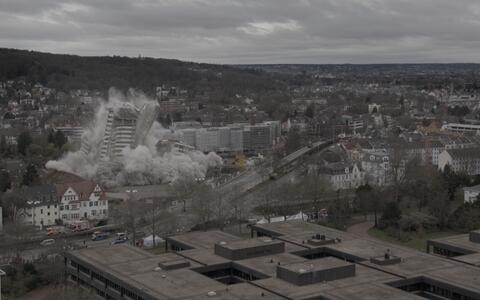 Eine weite, hohe Außenansicht einer Kleinstadt. Ein Gebäude in der Stadt steht mitten im Zusammensturz, um seinen Sockel steigt Rauch auf.