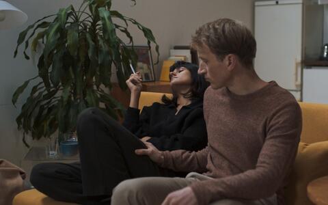 Eine Frau und ein Mann sitzen auf einem senfgelben Sofa. Die Frau racuht zurückgelehnt eine Zigarette, der Mann ist leicht nach vorn gebeugt und hat seine Hand auf ihrem Oberschenkel. Im Hintergrund ist eine Küchenzeile