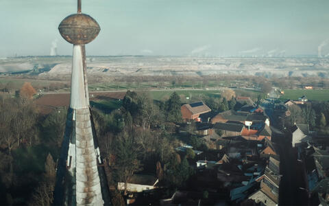 Im Vordergrund sieht man ein Dorf von oben und im HIntergrund einen Braunkohletagebau und Industrieschornsteine