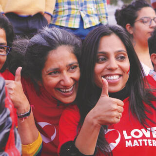 Drei Frauen mit roten Shirts mit der Aufschrift "Love Matters" stehen in einer Menschengruppe und halten die Daumen nach oben