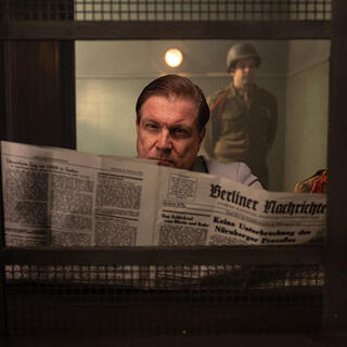 Ein Mann mit einer Zeitung schaut aus einem Fenster. Rechts im Hintergrund steht ein Soldat